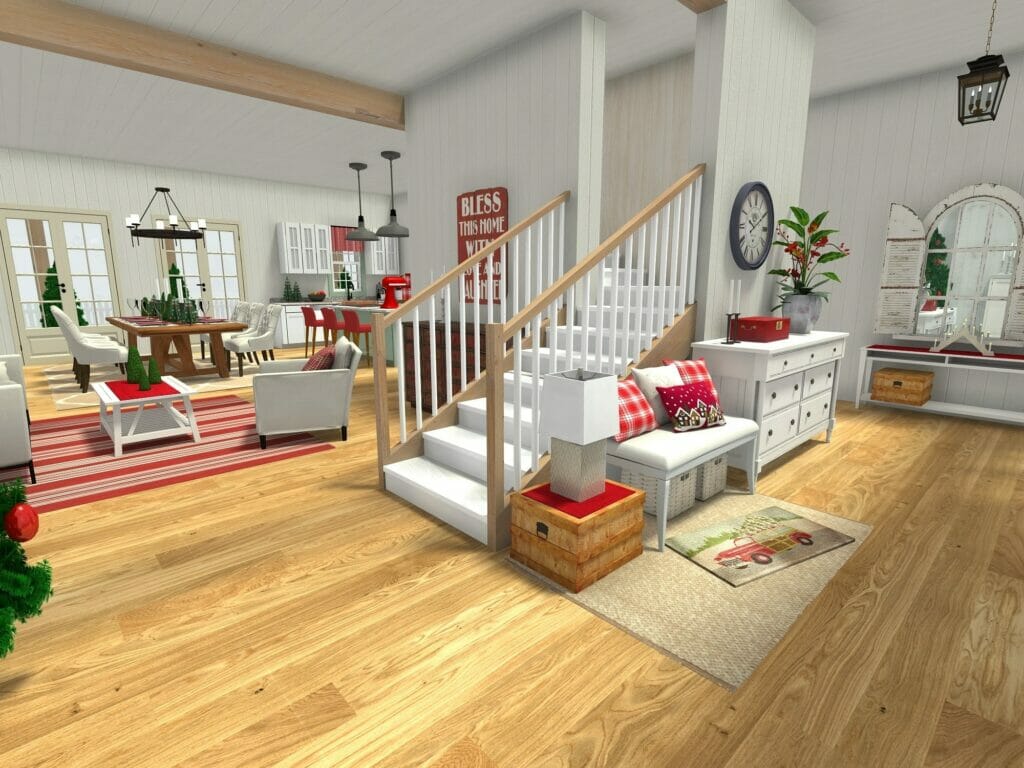 3D-rendered image of a cozy, holiday-themed open-concept living space with a staircase in the center. The room features light wood flooring, white paneled walls, and festive red and green accents. A dining area with a rustic table and a kitchen with pendant lighting are visible in the background. The foreground includes a cushioned bench with holiday pillows, a dresser with decor, and a “Bless This Home With Love” sign on the wall.