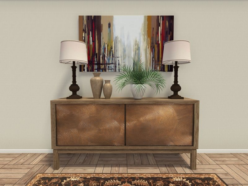 Stylish entryway or living room vignette featuring a wooden console table with a circular woodgrain pattern on the doors. The tabletop is decorated with two matching table lamps, a pair of ceramic vases, and a potted green plant. A colorful abstract cityscape painting hangs on the wall above, and the space is anchored by a patterned rug and herringbone wood flooring.