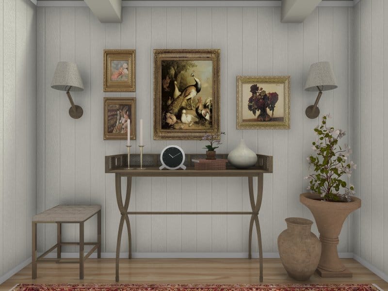 Elegant vignette of a traditional-style interior corner featuring a slim console table with decorative objects like candles, a clock, and vases. The wall is adorned with classic framed paintings and flanked by two wall-mounted sconces. A cushioned bench sits on the left, and on the right, two rustic floor vases and a large planter with blooming branches add charm to the neutral, paneled wall backdrop.