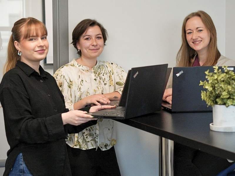RoomSketcher's customer support team standing around a tall black table, each working on a laptop and smiling at the camera. 
