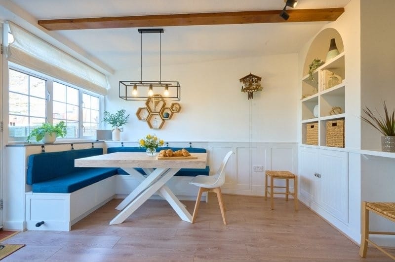 Bright dining nook with a wooden table, white cross-leg base, and built-in bench seating with blue cushions. Large windows let in natural light, while open shelving, plants, and honeycomb wall decor add warmth and character.