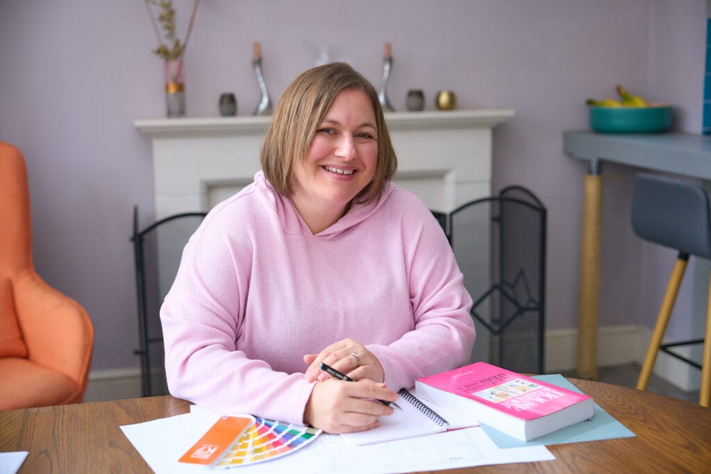 Interior designer Debbie Bryant in a pink hoodie sitting at a wooden table with design materials, including a color swatch, notebook, and a bright pink book. The background shows a cozy room with lavender walls, a fireplace, a modern bar counter, and an orange armchair. She appears to be working on an interior design project.