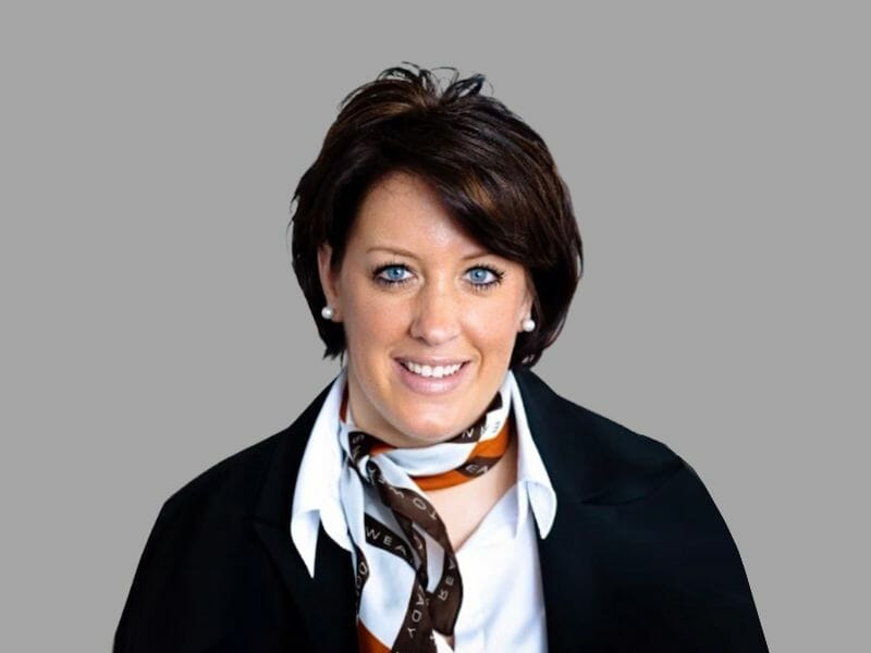 Professional portrait of real estate agent Manuela Schroeder with short dark brown hair, wearing a black blazer, white blouse, and a patterned scarf. She is smiling and posed in front of a plain gray background.