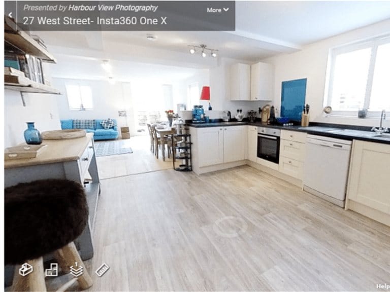 3D virtual tour view of a bright open-plan kitchen and dining area captured with Matterport. The kitchen features white cabinets, black countertops, and modern appliances, with a dining table and blue sofa visible in the background. Light wood flooring runs throughout the space.