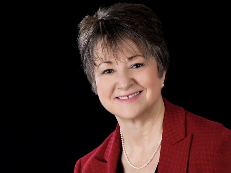 Robin Tschumper Interior Designer with short brown hair wearing a red blazer, black top, and a pearl necklace. The background is solid black, creating a formal and polished look.
